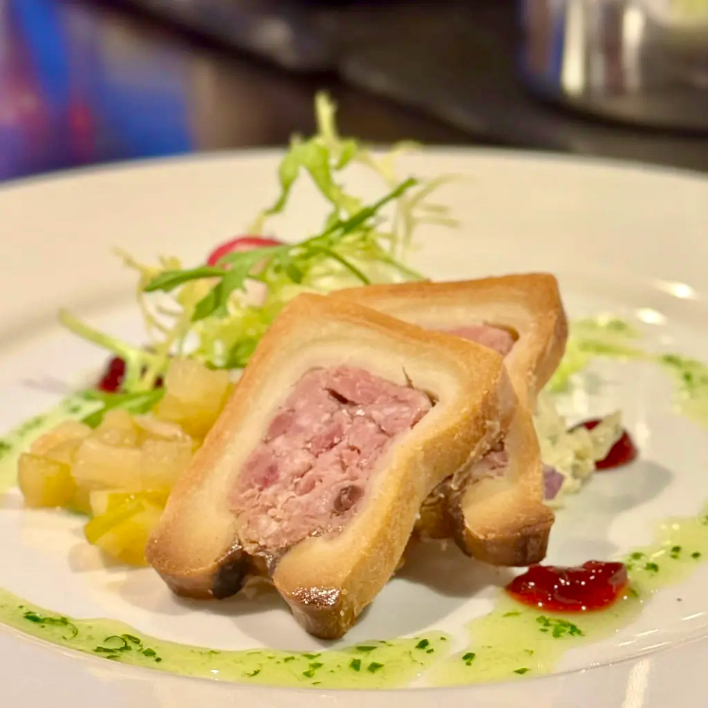 Slices of pâté en croûte with frisée, pickled vegetables, and a herb oil drizzle