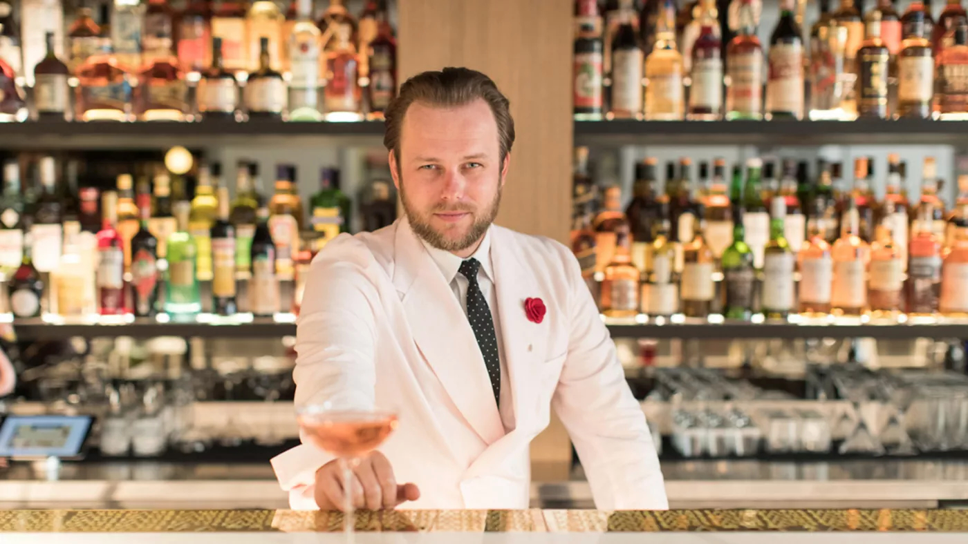 A bartender in a cream jacket with a red rose, sliding a cocktail across a polished bar