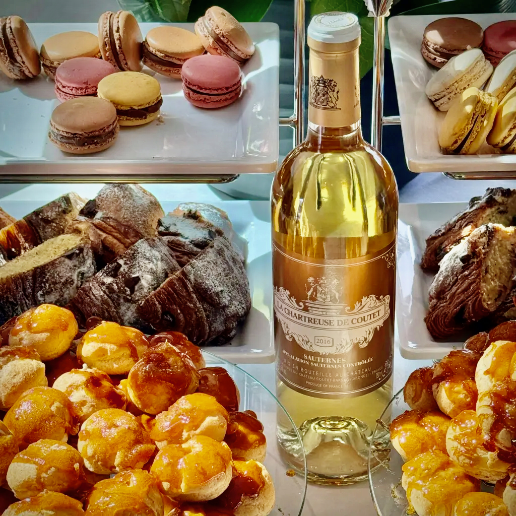 Tiered trays of macarons, chocolate croissants, and caramel-glazed choux buns beside a bottle of Sauternes
